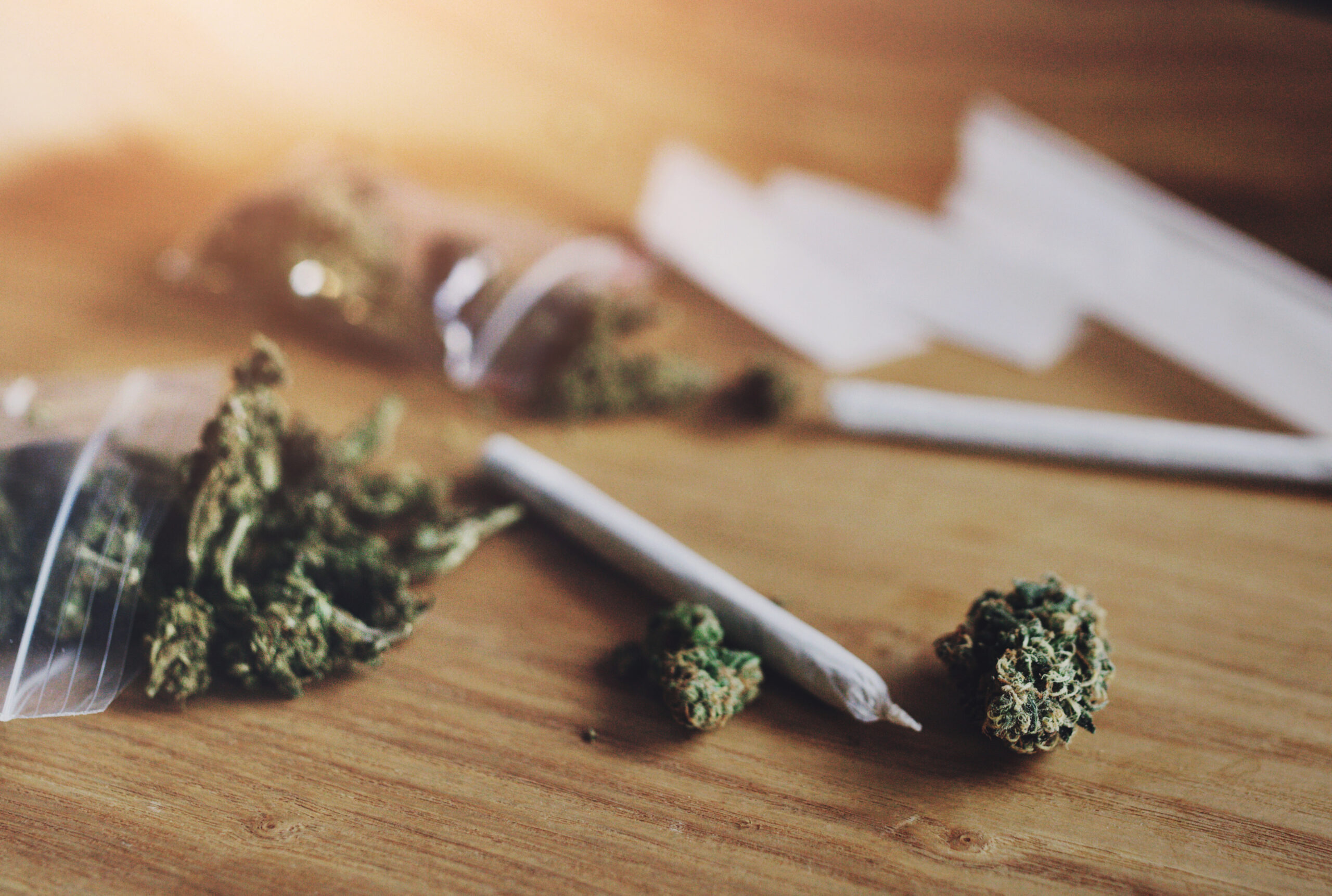 Close-up of cannabis buds, rolled joints, and rolling papers on a wooden surface, with a warm, natural light.